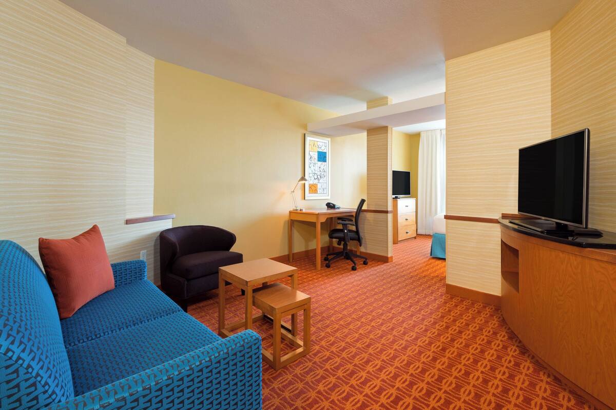 A hotel room with a blue sofa, a small wooden table, a desk with a chair, a television, and a patterned carpet.
