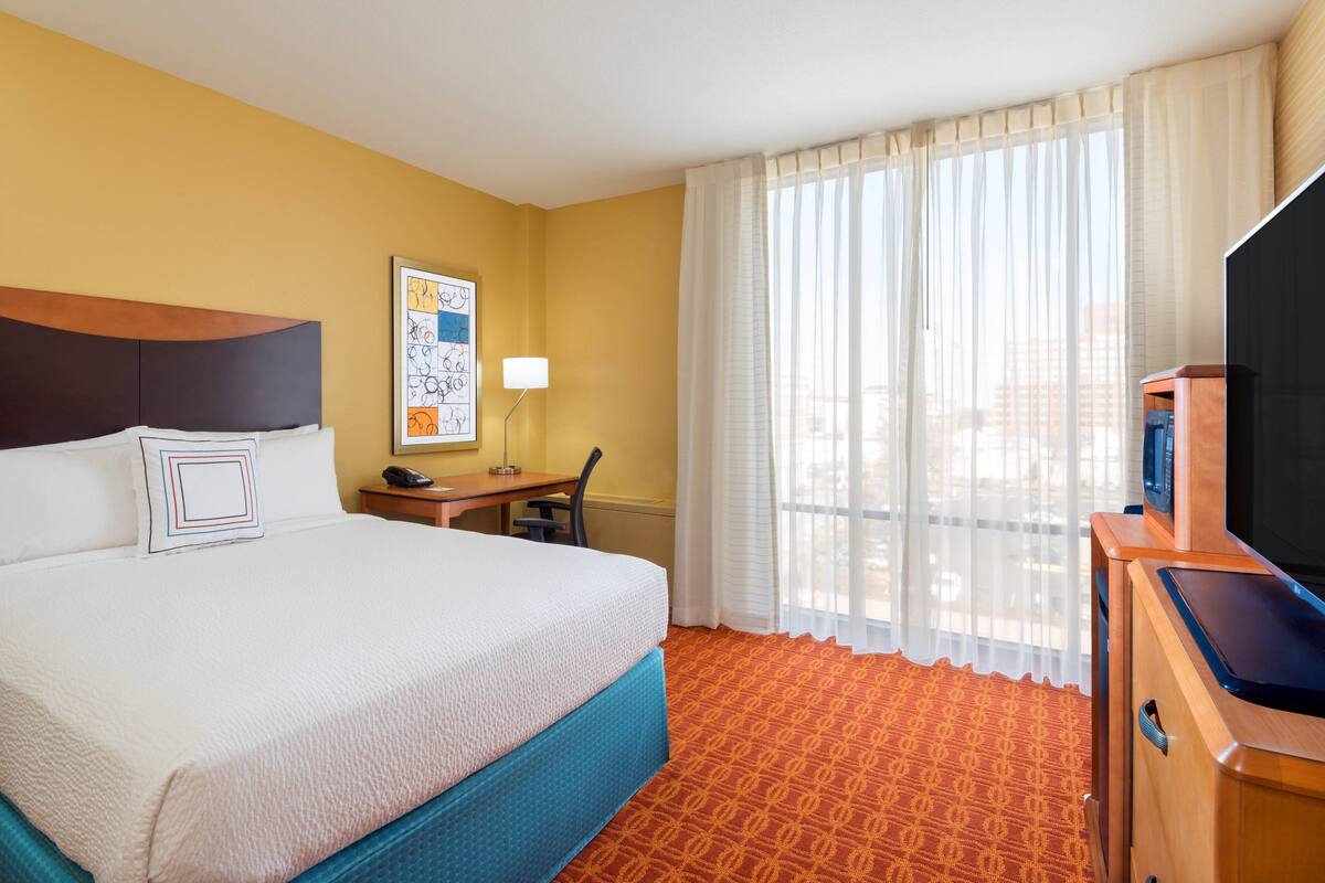 A hotel room with a large bed, a desk with a lamp, a television, and a window with sheer curtains.