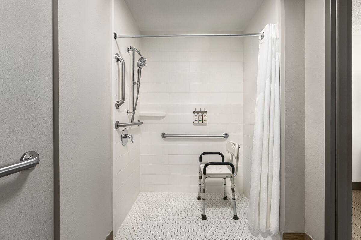 A small, accessible shower room with a white tiled floor, a grab bar, a shower chair, and a handheld showerhead.
