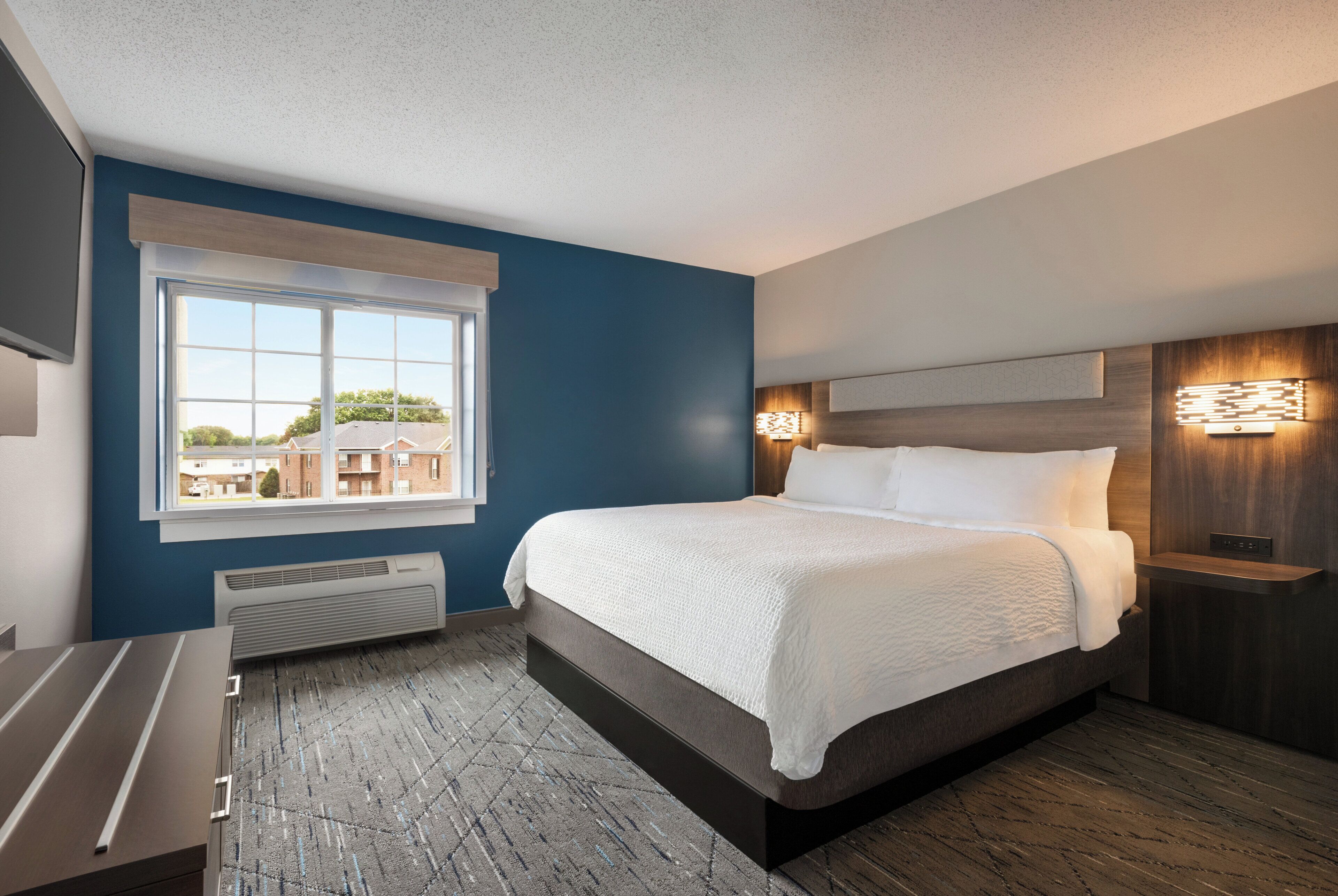 Una habitación de hotel moderna con una cama grande, una ventana con persianas, un calentador integrado y una mesita de noche con lámpara.