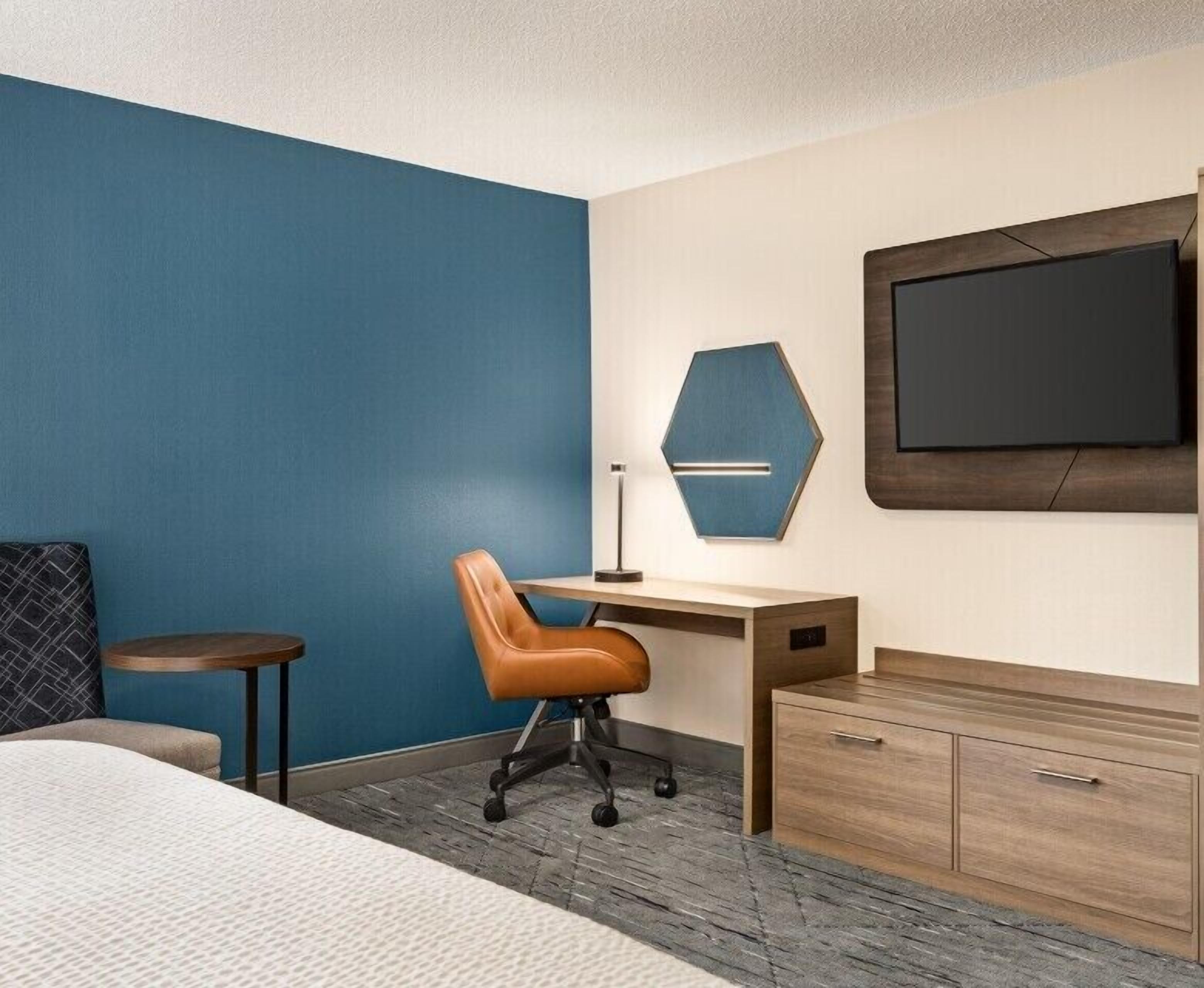 Een hotelkamer met een bed, bureau, stoel, klein tafeltje, lamp, televisie en een houten kast.