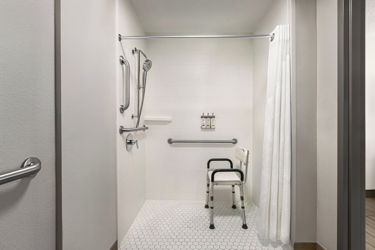 Een badkamer met witte betegelde wanden, een witte vloer, een douche met een glazen deur, een witte stoel en een handgreep.
