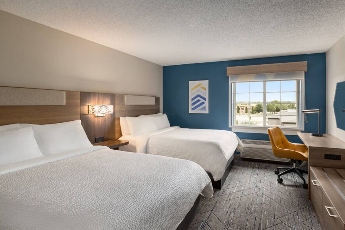 A hotel room with two beds, a desk, a chair, a window with a view, and a wall-mounted artwork.
