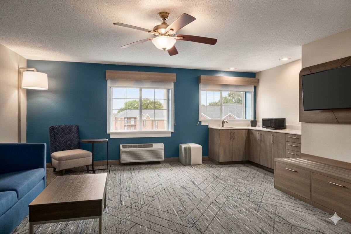 A modern room with a ceiling fan, a sofa, a coffee table, a small table, a TV, a window, and a kitchen area.
