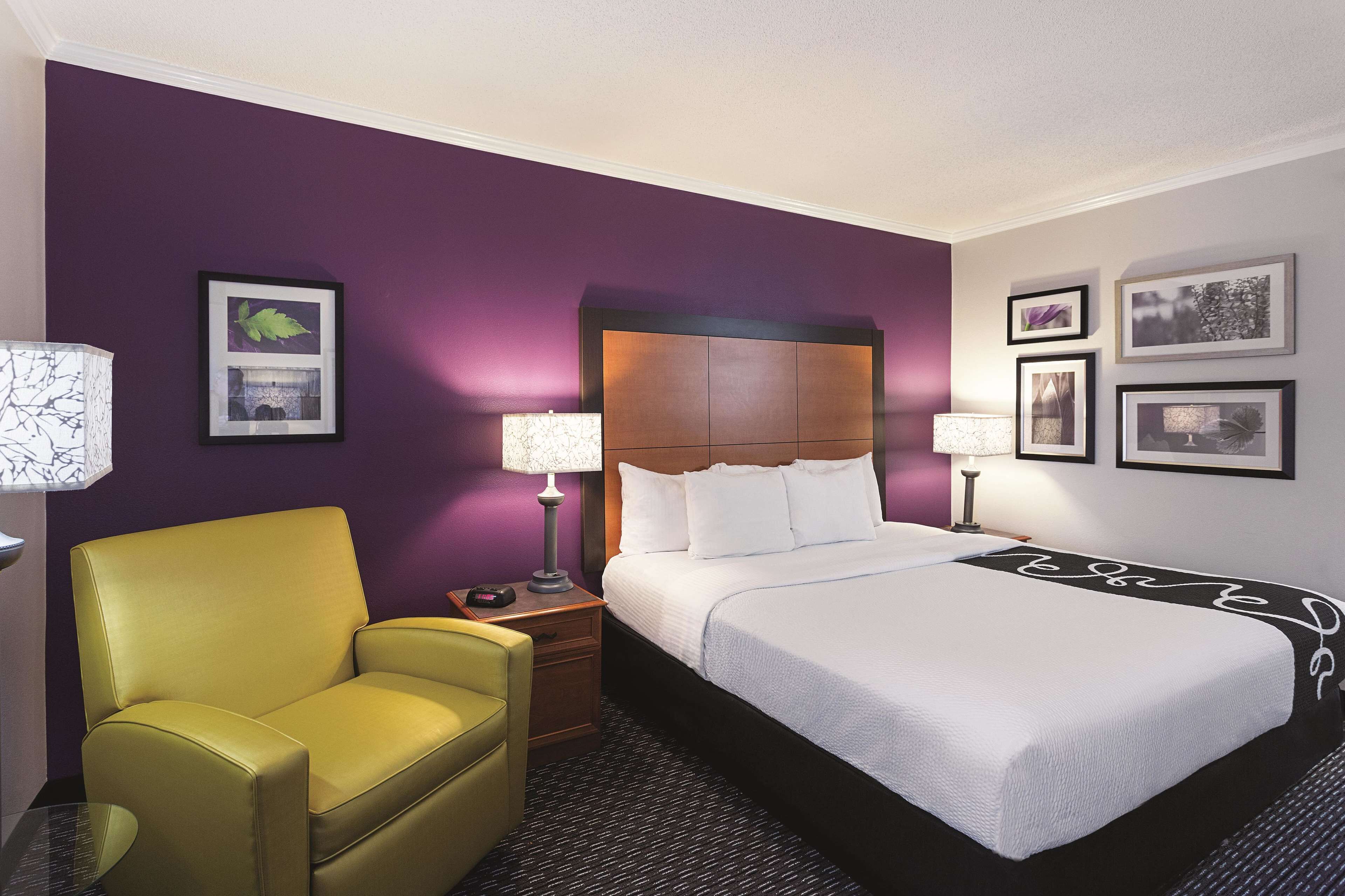 Quarto de hotel com uma cama grande, uma poltrona amarela e uma mesa de cabeceira com abajur.