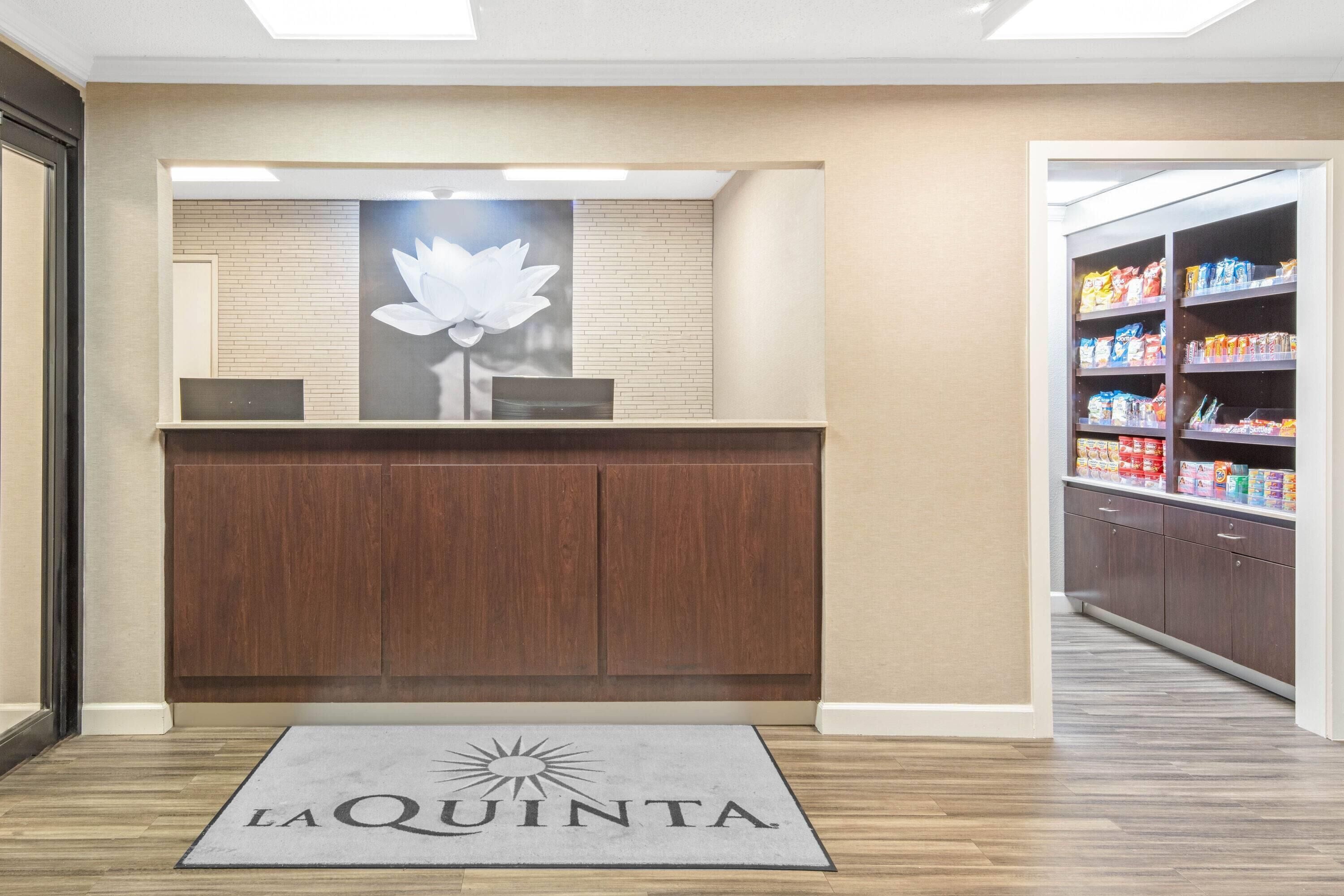 A reception area with a wooden counter, a large floral artwork, and a 'La Quinta' doormat.