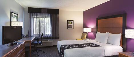 A hotel room with a bed, a desk with a television, a chair, a window with curtains, and a wall-mounted lamp.