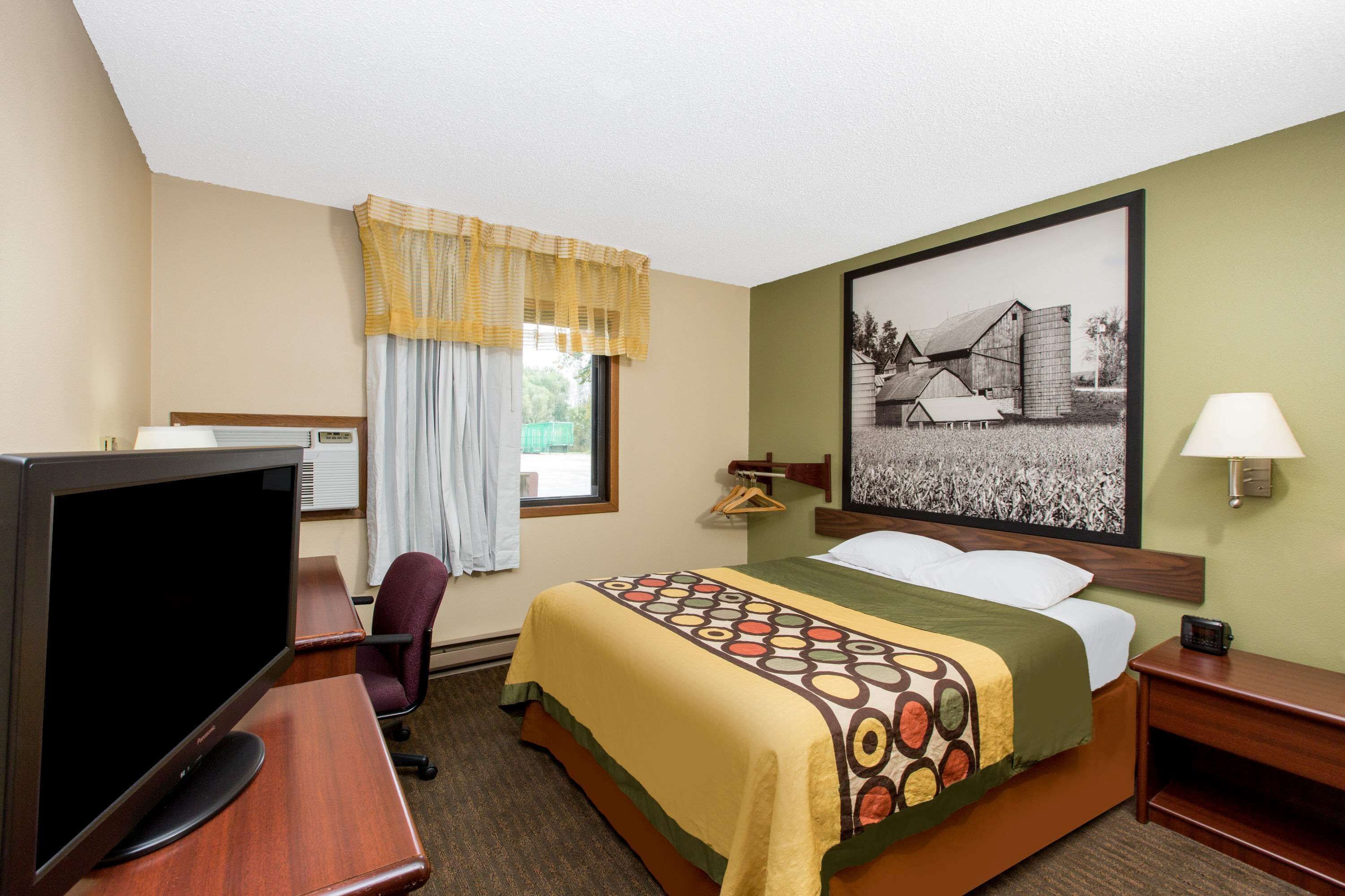 A hotel room with a bed, a desk with a computer, a chair, a window with curtains, a wall-mounted lamp, and a framed picture on the wall.