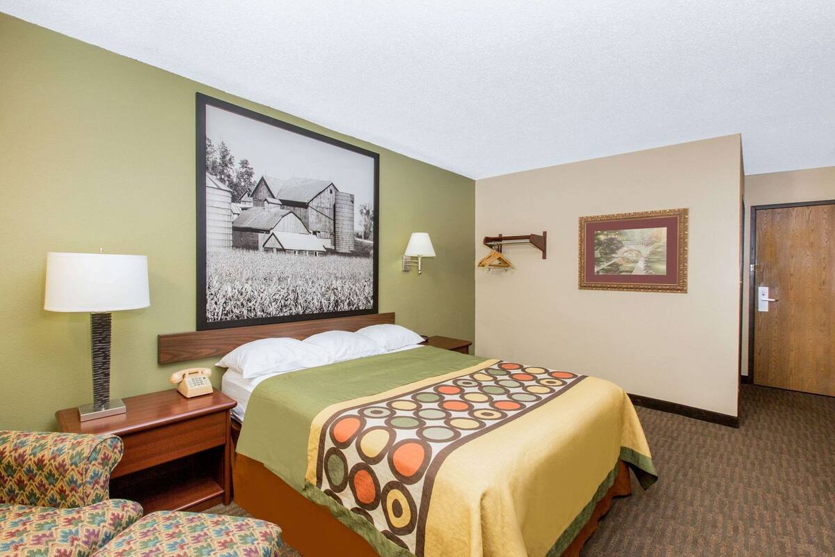 Une chambre d’hôtel avec un lit, deux lampes de chevet, un téléphone, une chaise, un bureau, un tableau encadré au mur et une porte.