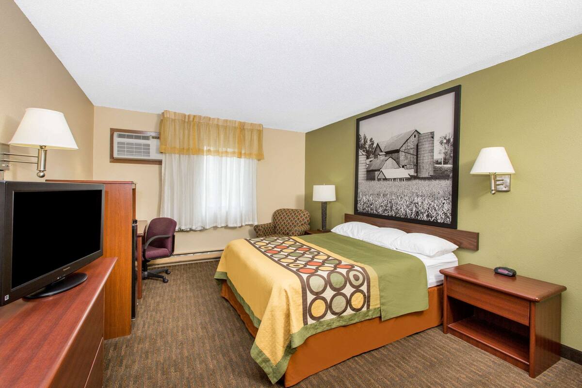 Une chambre d’hôtel comprenant un lit, un bureau avec une télévision, une chaise, une table de chevet et une lampe fixée au mur.