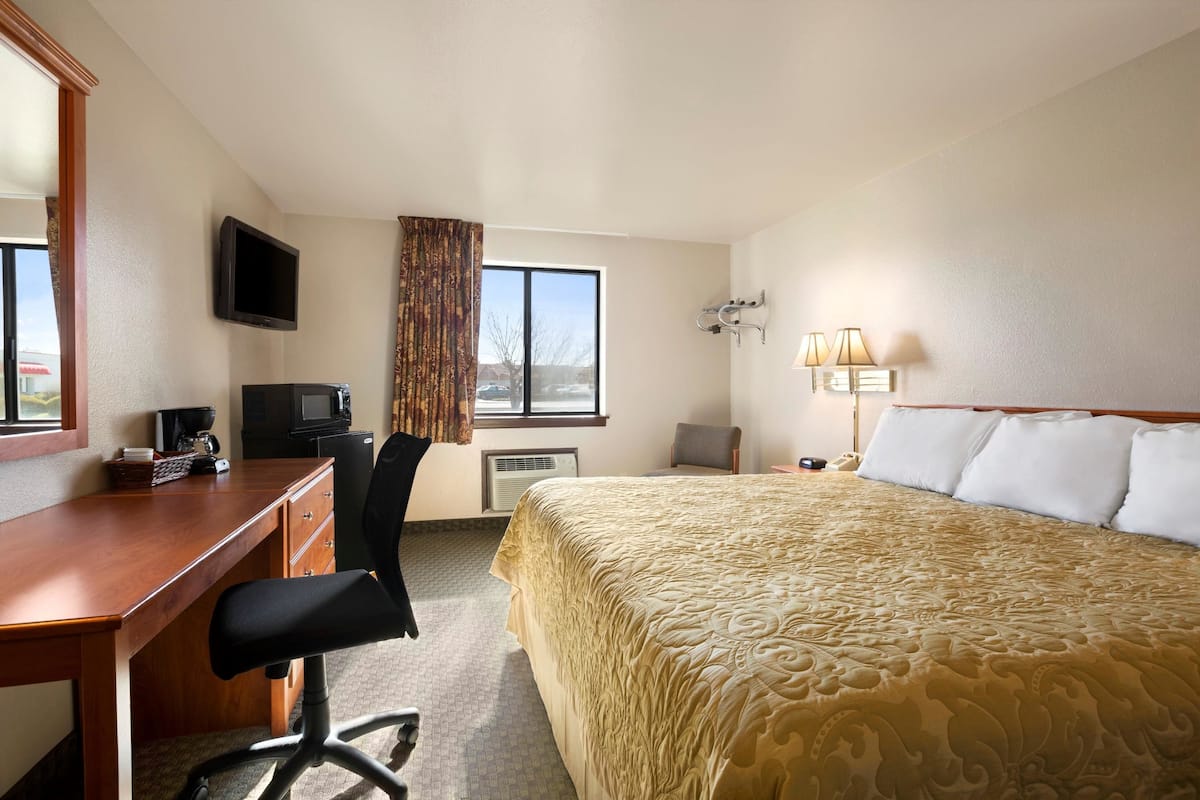 A hotel room with a wooden desk, a black office chair, a bed with a gold patterned bedspread, a television mounted on the wall, a window with curtains, and a wall-mounted lamp.
