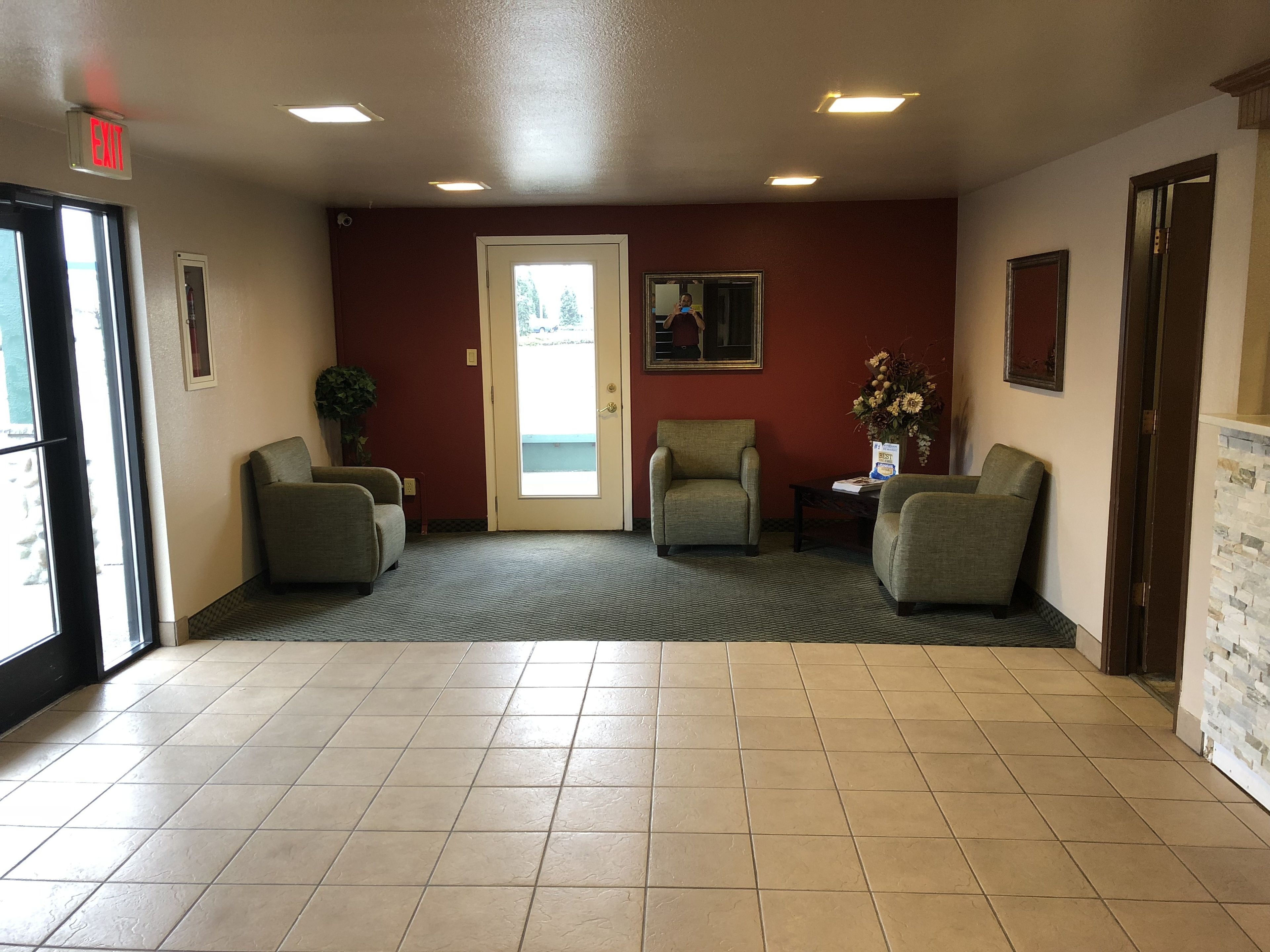 A reception area with a red wall, two armchairs, a small table with a vase, and a door with an exit sign.