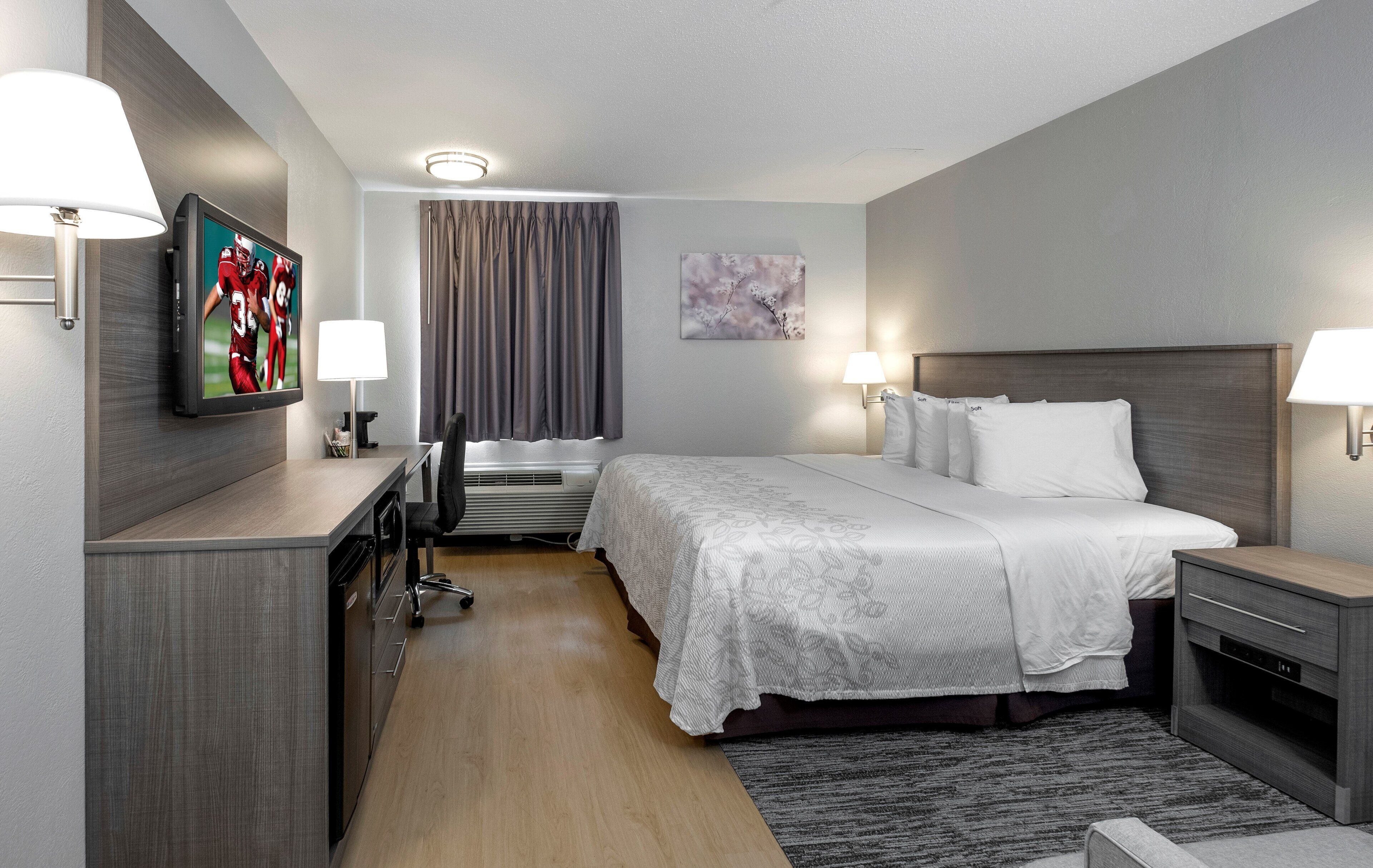 Une chambre d’hôtel comprenant un lit, des tables de chevet, un bureau, une chaise, une télévision, une lampe, un tableau et une fenêtre avec des rideaux.