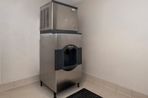 Un refrigerador comercial de acero inoxidable con dispensador de agua, ubicado en una esquina con piso de baldosas.