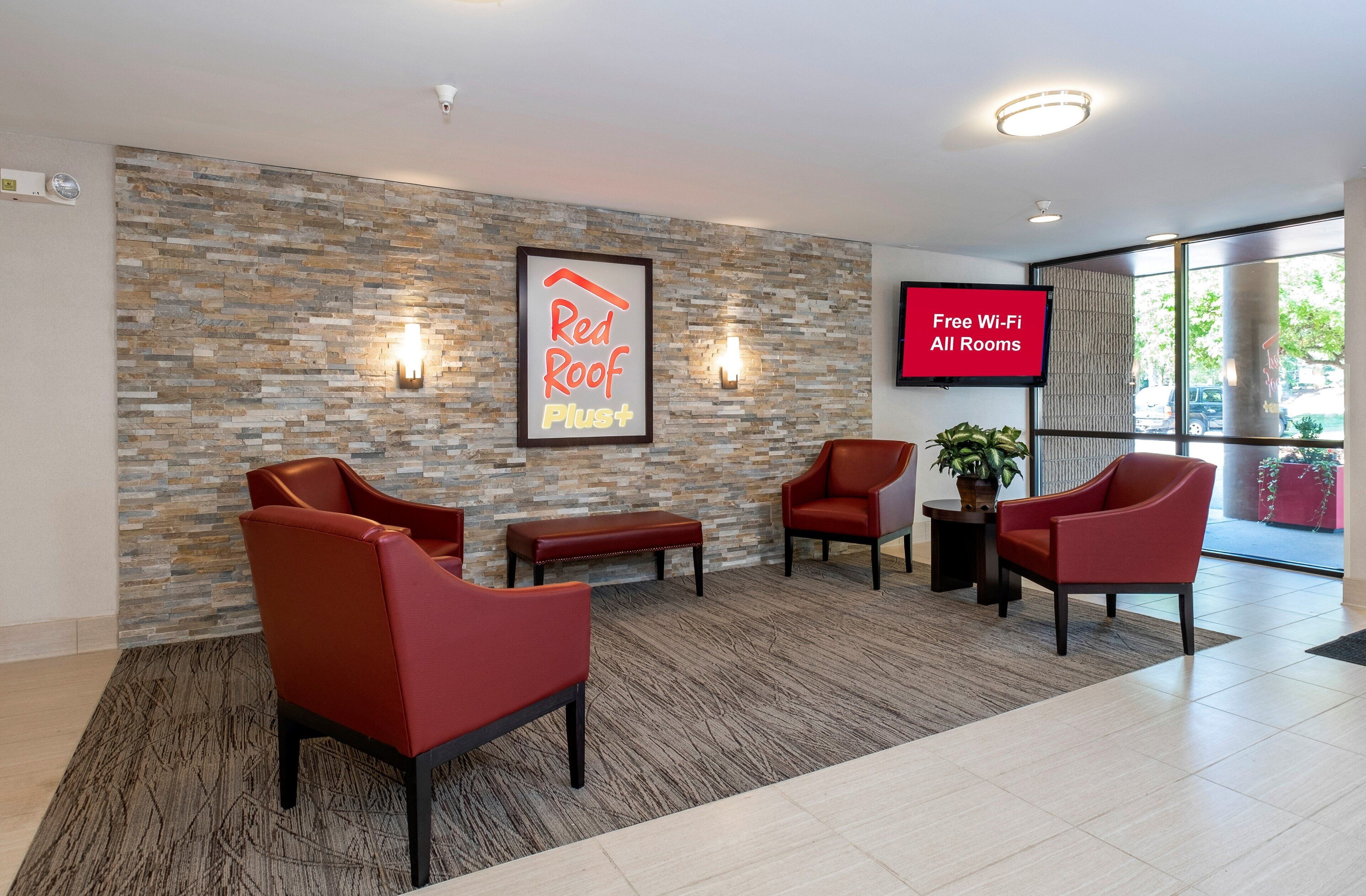 A hotel lobby with a stone wall, red chairs, a bench, a TV screen advertising free Wi-Fi, and a framed sign for Red Roof Plus.