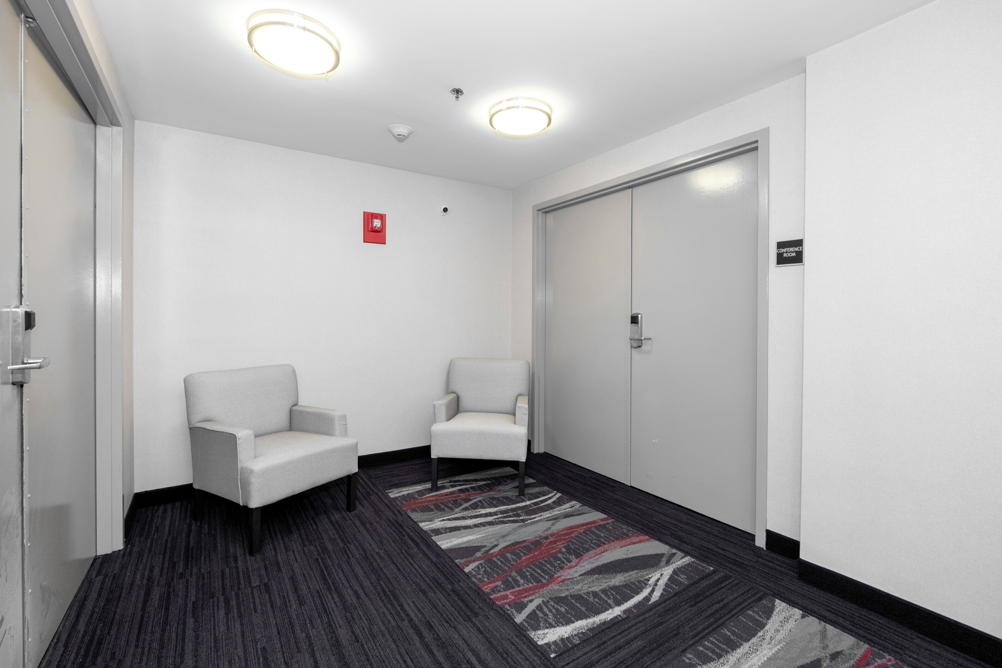 A hallway with two white armchairs, a carpeted floor, a door labeled 'Conference Room', and ceiling lights.