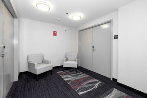 Un pasillo con dos sillones blancos, piso alfombrado, una puerta con la inscripción 'Sala de Conferencias' y luces en el techo.