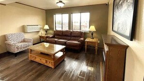 A living room with a brown leather sofa, patterned armchair, and a wooden coffee table. There are two windows with a view of a parking lot and a building. - FairBridge Inn & Suites Moscow/Pullman (Moscow)