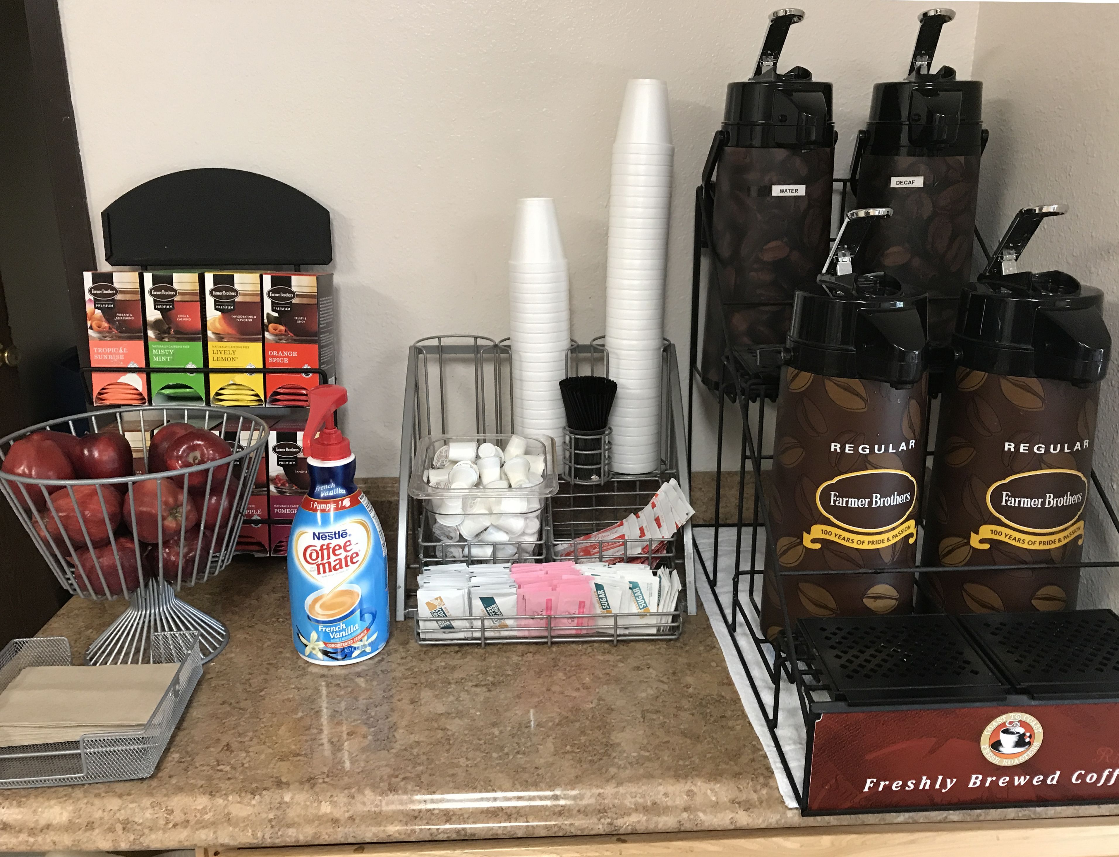 A coffee station with coffee makers, disposable cups, and a basket of apples.