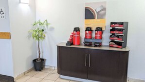 Een receptiezone met een potplant, een koffiehoek met rode dispensers, en een bord met de tekst 'RISE & SHINE'.