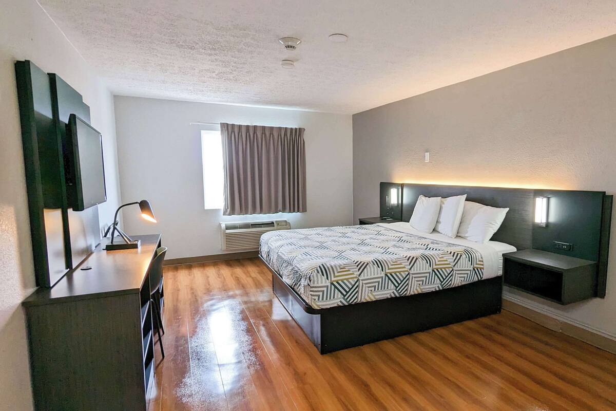 A modern hotel room with a wooden floor, a large bed with patterned bedding, a desk with a chair, a flat-screen TV, and a window with curtains.