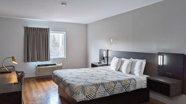 A modern hotel room with a large bed, bedside tables, a desk with a lamp, a window with curtains, and a wall-mounted light fixture. - Motel 6 Framingham, MA - Boston West (Framingham)