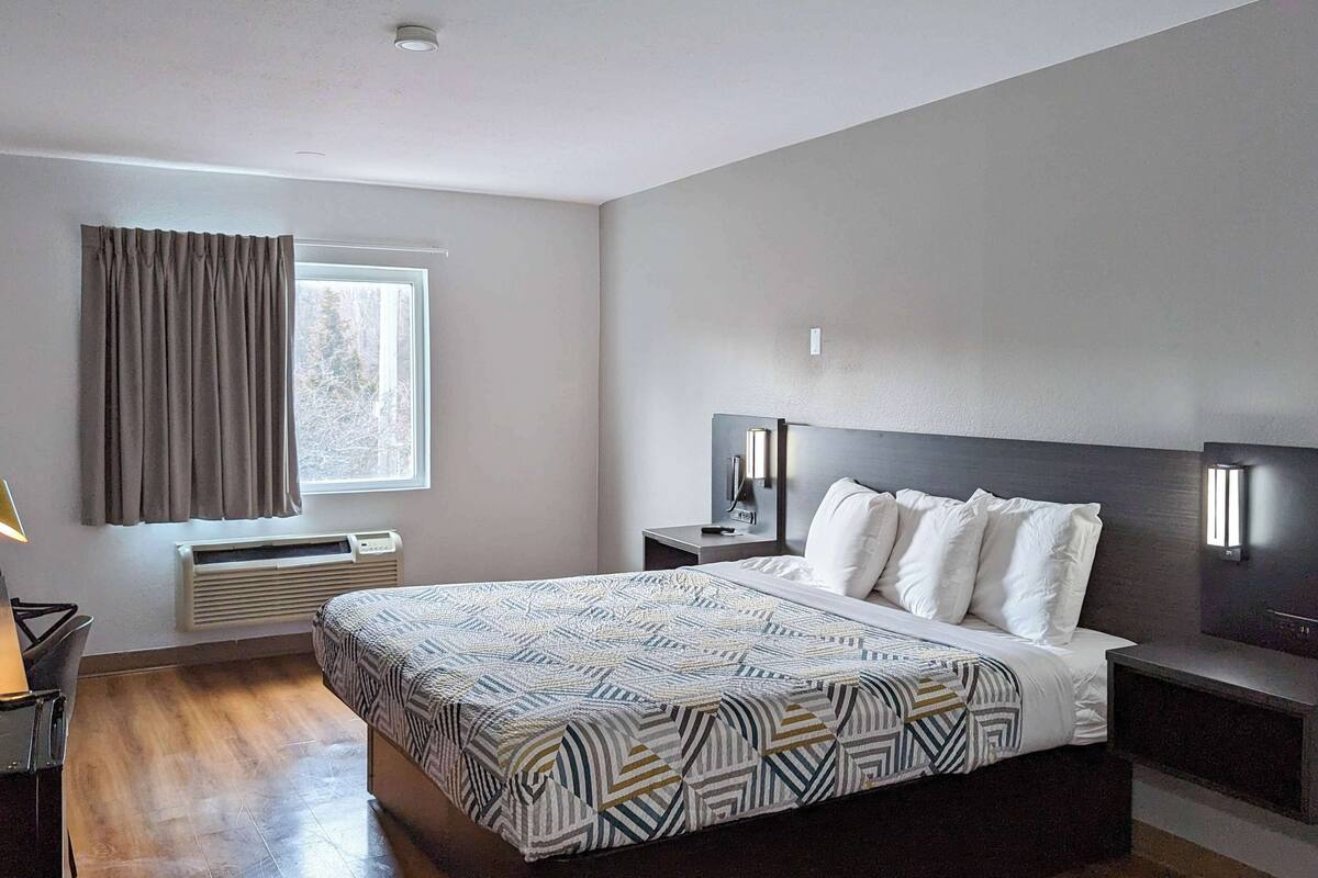 A modern hotel room with a large bed, bedside tables, a desk with a lamp, a window with curtains, and a wall-mounted light fixture.