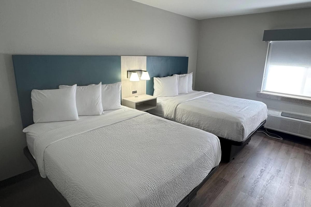 Habitación de hotel con dos camas, una mesita de noche con lámpara, una ventana con persianas y un aire acondicionado de pared.