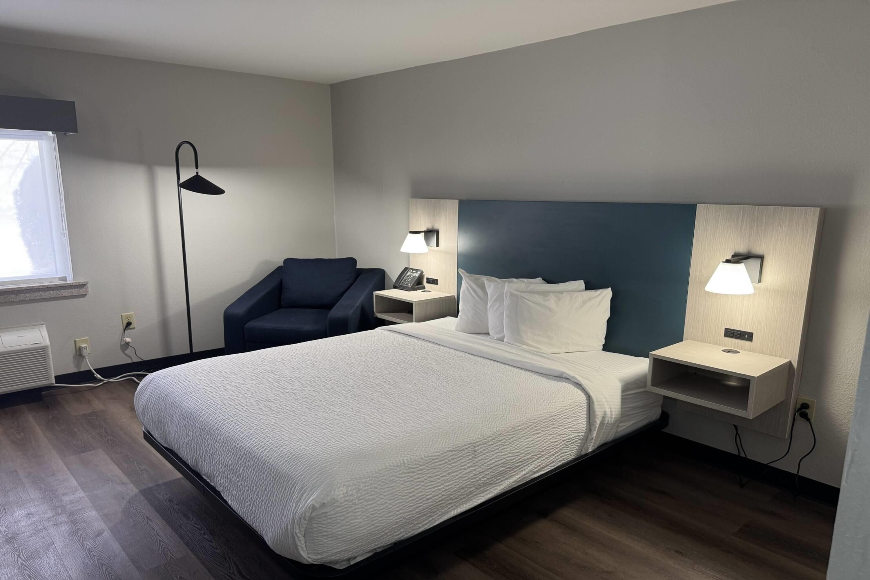 Una habitación de hotel con cama, mesitas de noche, una silla, un escritorio, una lámpara y una ventana.