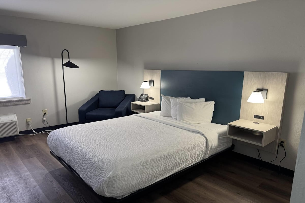 Una habitación de hotel con cama, mesitas de noche, una silla, un escritorio, una lámpara y una ventana.