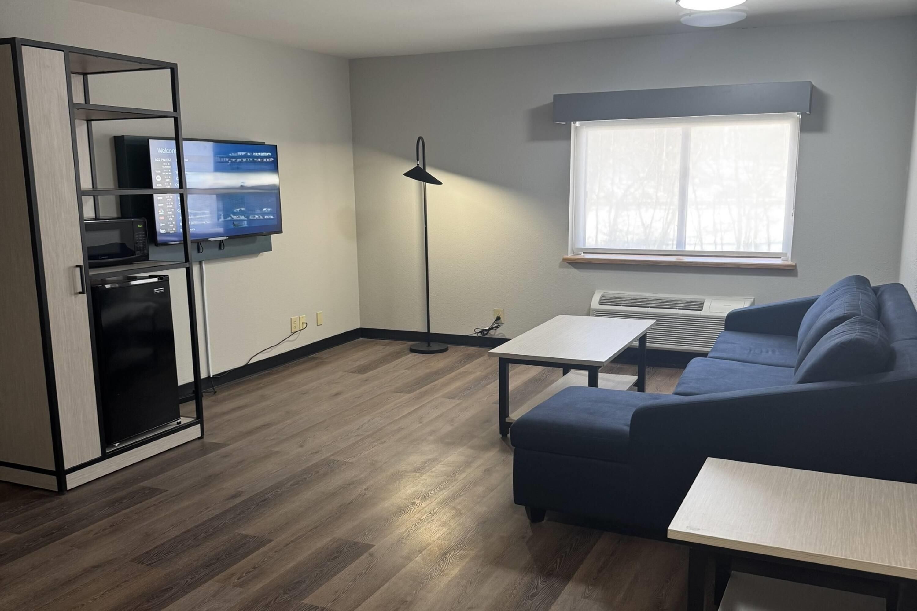 Una sala moderna con un sofá, una mesa de centro, un televisor montado en la pared, una lámpara de pie y una cocina integrada.