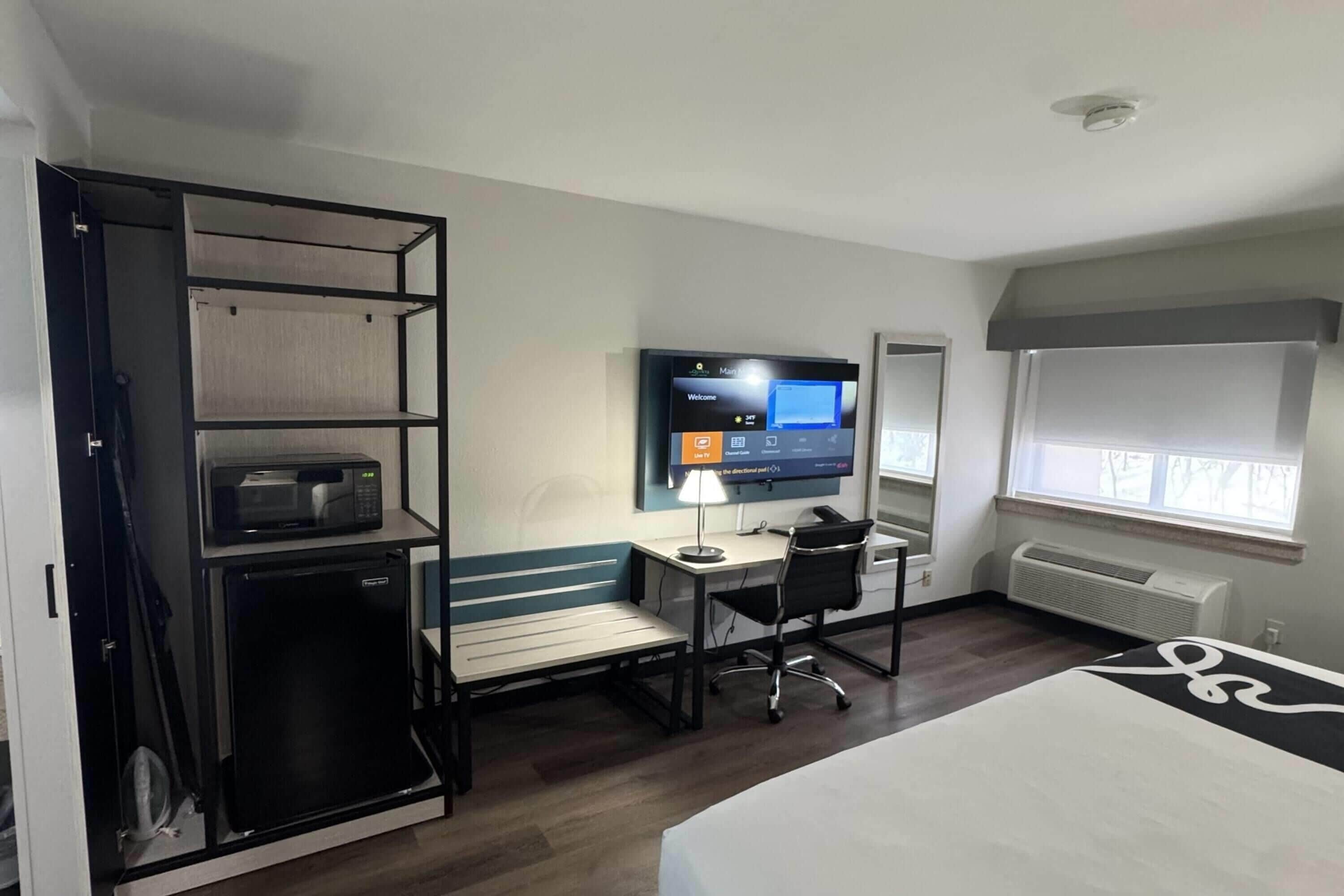 Habitación de hotel con cama, escritorio con computadora, televisión, microondas, refrigerador pequeño y ventana con persianas.