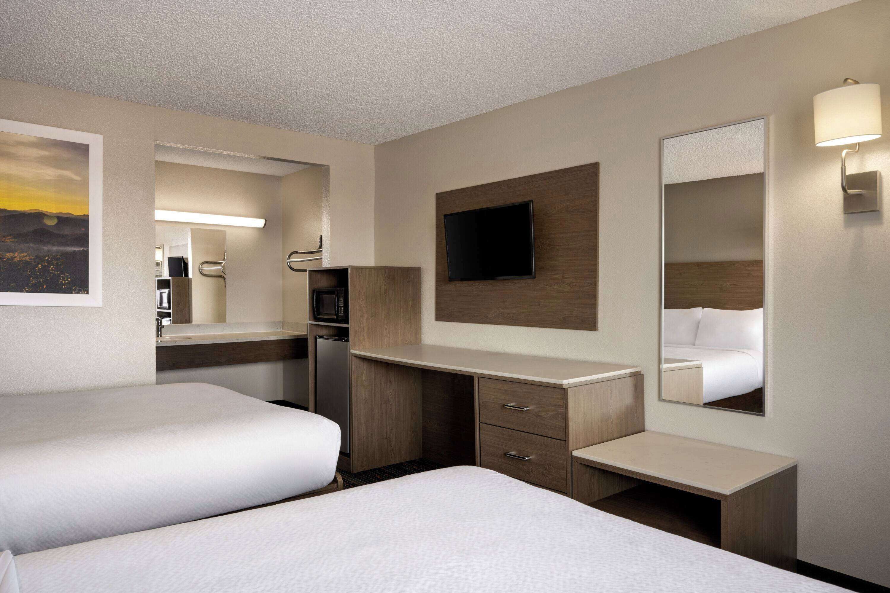 A hotel room with two beds, a desk, a flat-screen TV, a mirror, and a wall-mounted lamp.