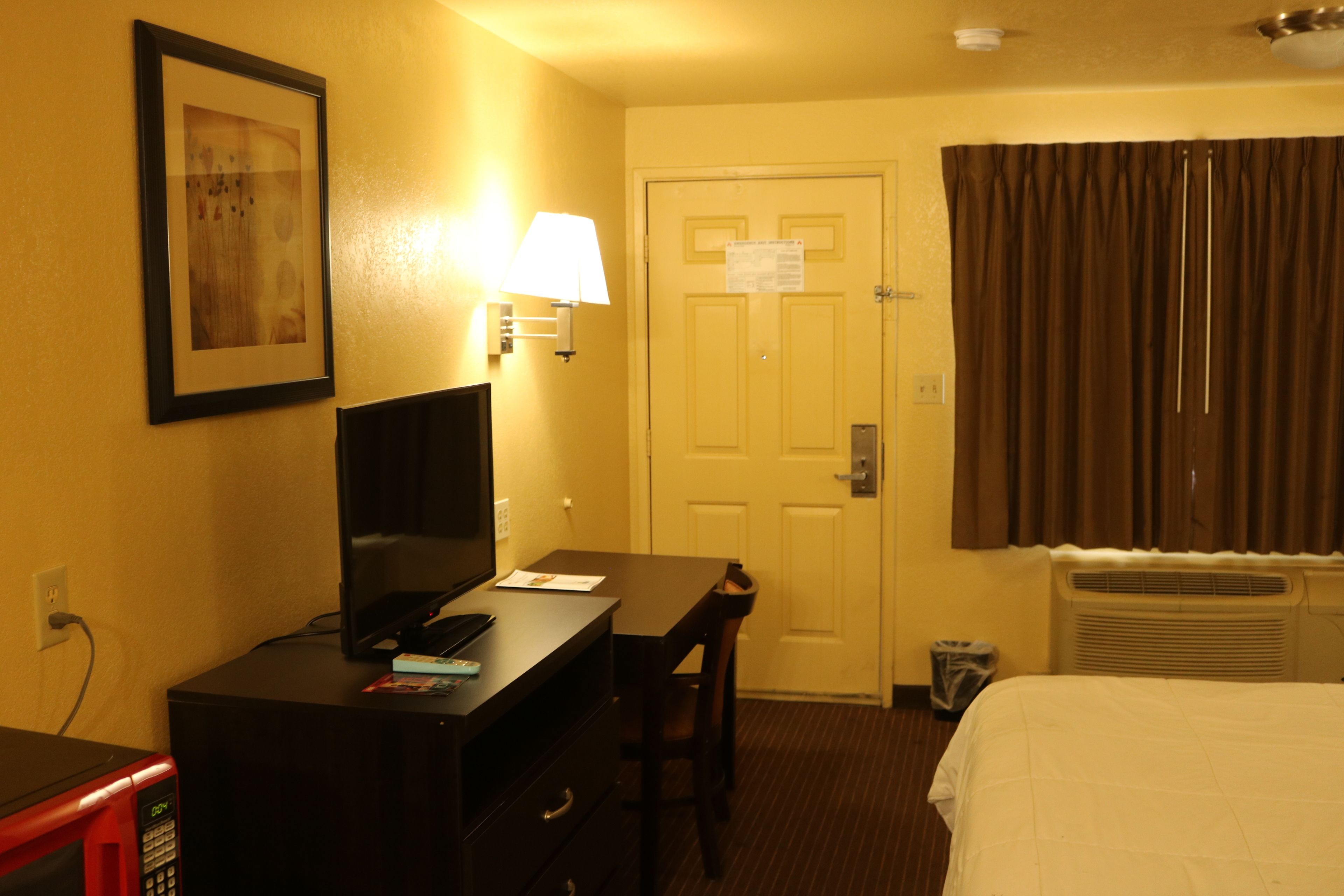 A hotel room with a desk, chair, bed, television, lamp, and a door with a sign.
