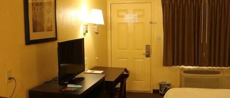 A hotel room with a desk, chair, bed, television, lamp, and a door with a sign.