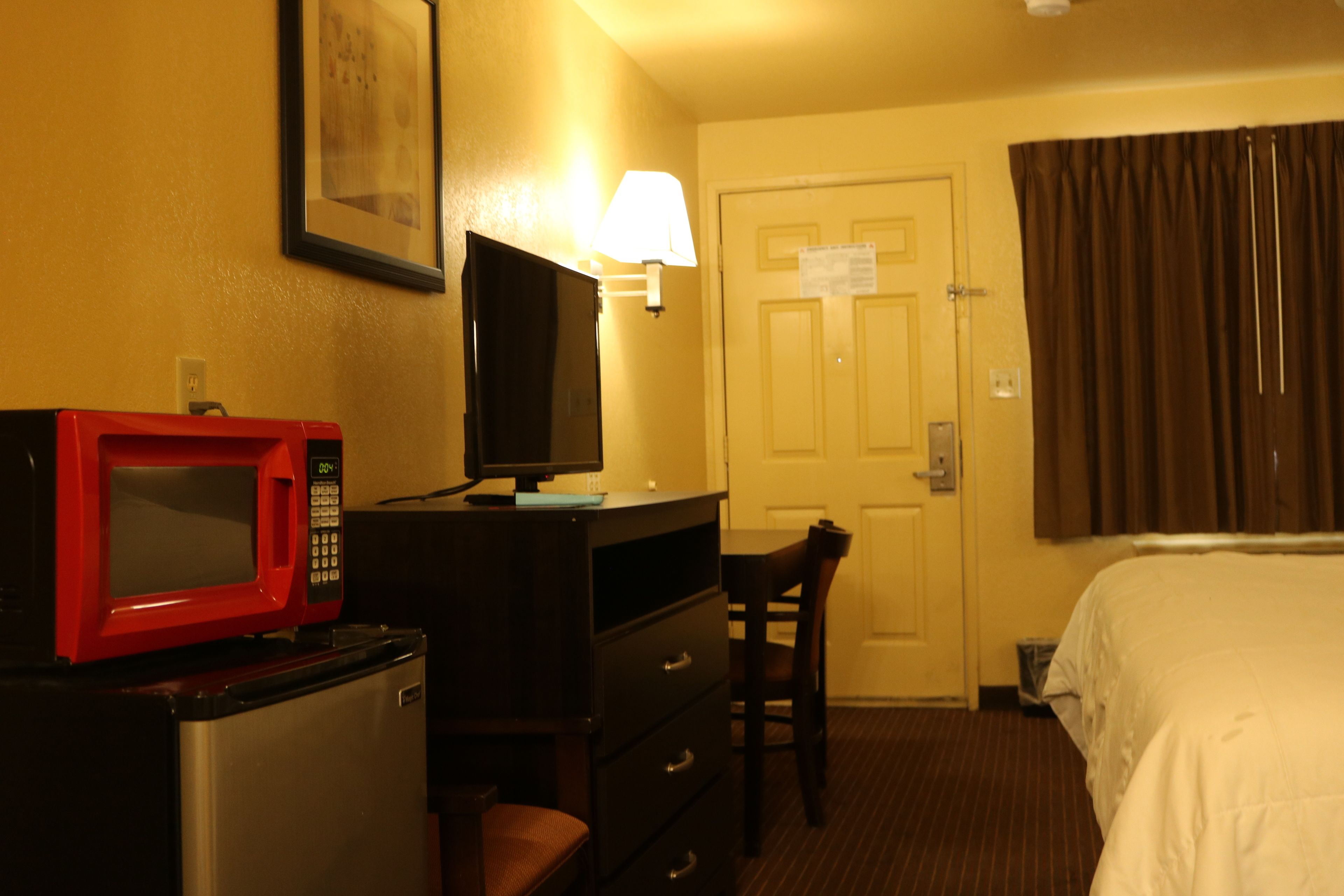 Una habitación de hotel con una cama, un escritorio con computadora y un microondas rojo, una silla, una lámpara y una puerta con un aviso.