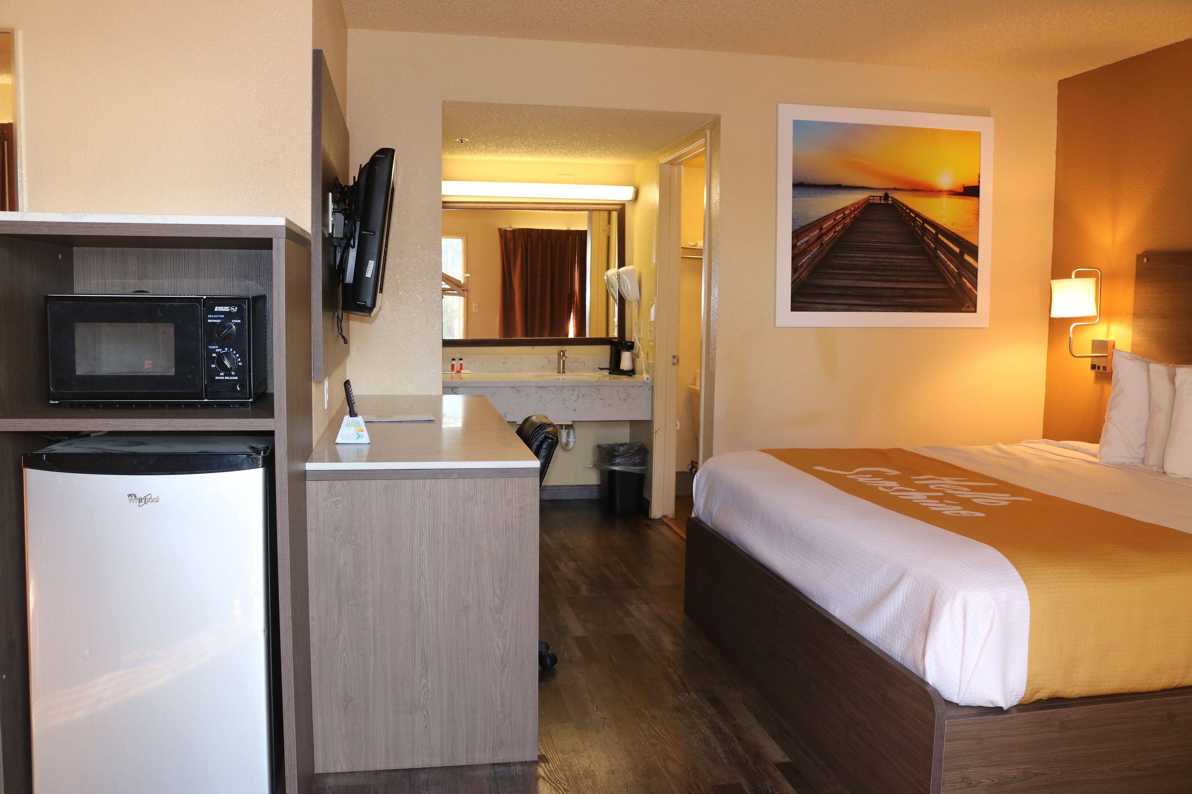 Une chambre d’hôtel comprenant un lit, un bureau avec un four à micro-ondes, un petit réfrigérateur, une salle de bain avec douche et un tableau représentant un coucher de soleil sur une jetée.