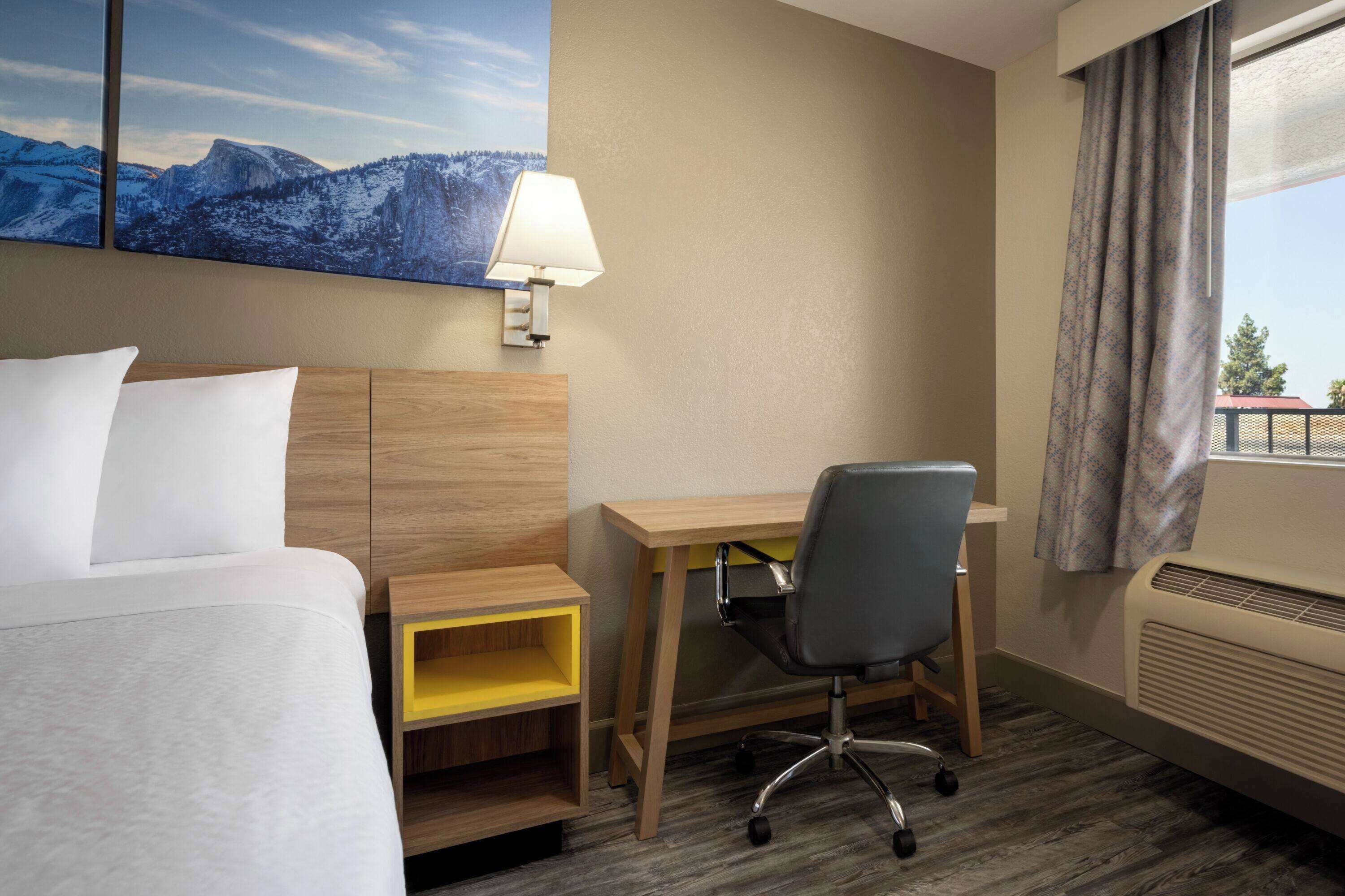 A hotel room with a bed, desk, chair, and a large window with curtains.