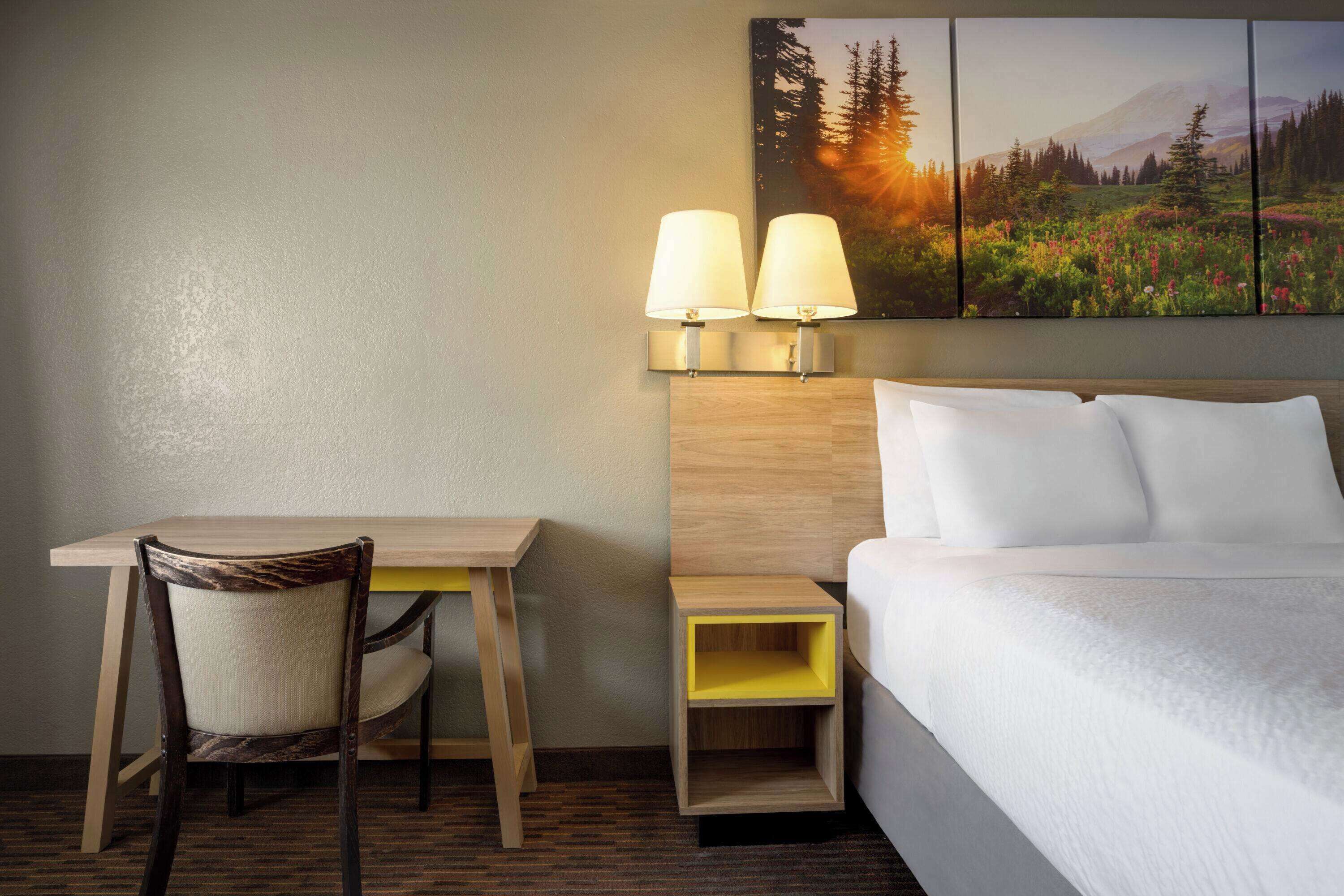 A hotel room with a bed, a desk with a chair, two wall-mounted lamps, and a large landscape painting above the bed.