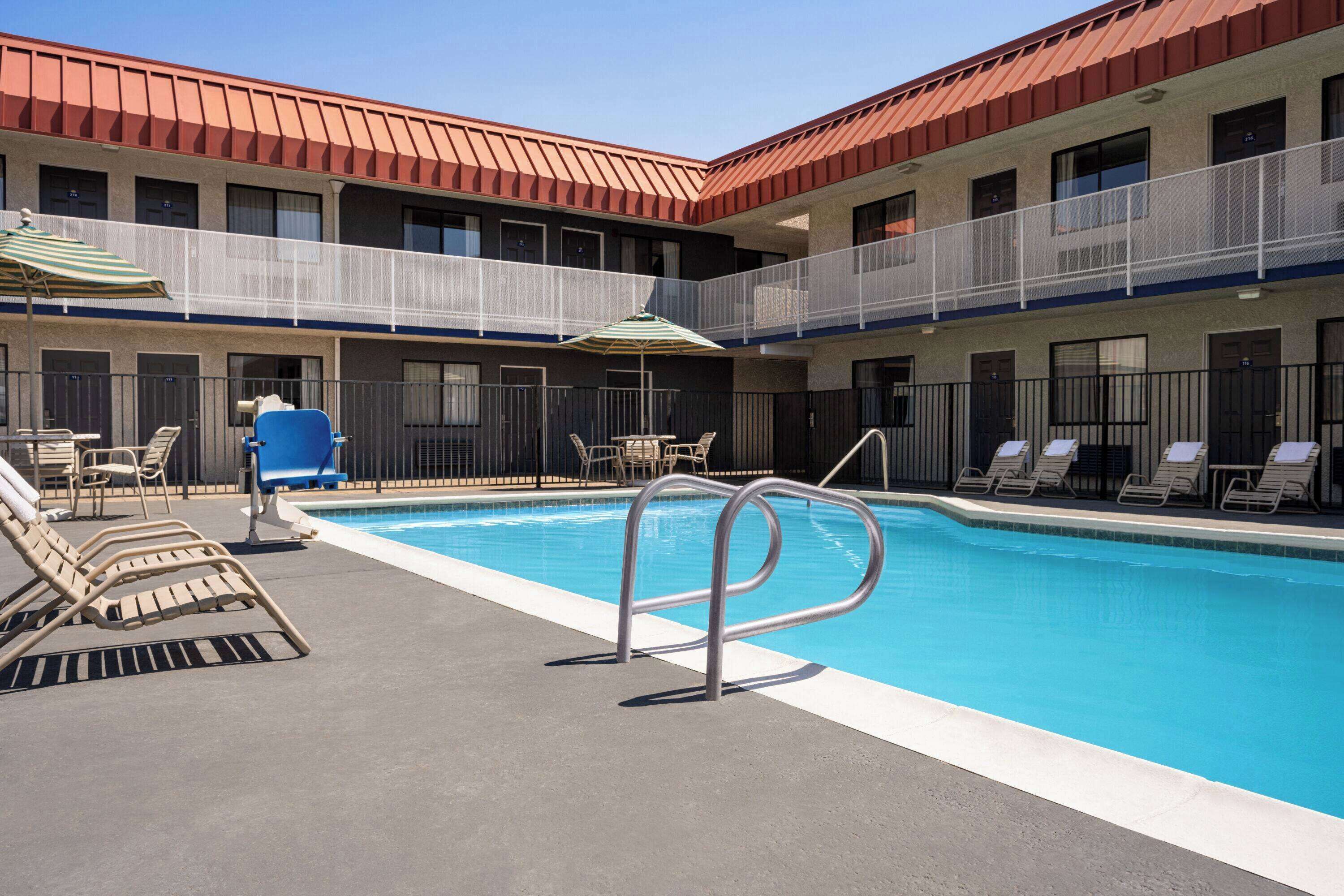 A hotel pool area with lounge chairs, a pool with a metal safety rail, and a covered seating area with umbrellas.