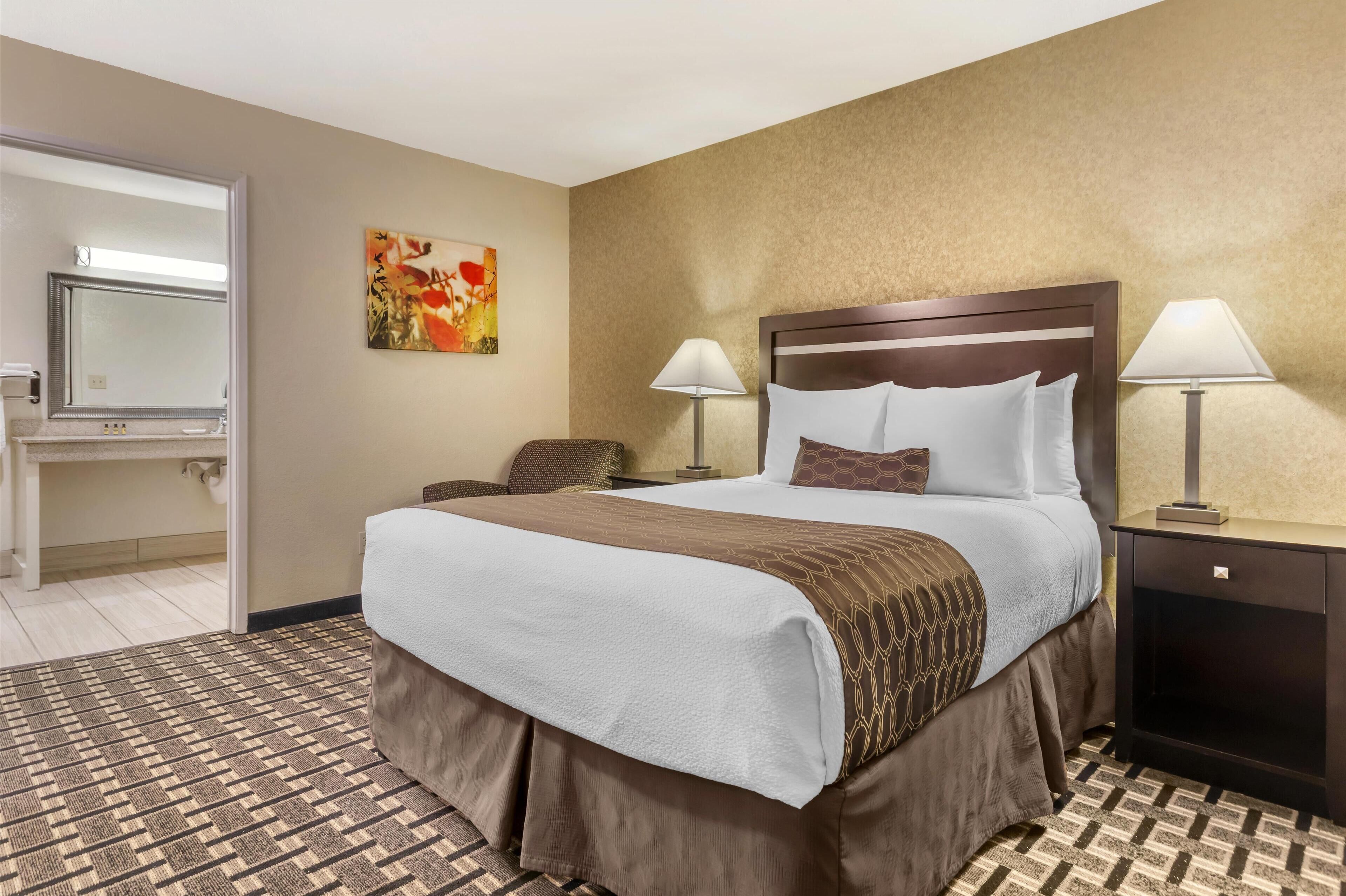 A hotel room with a large bed, two bedside lamps, a headboard, a small table, a chair, a mirror, and a patterned rug.
