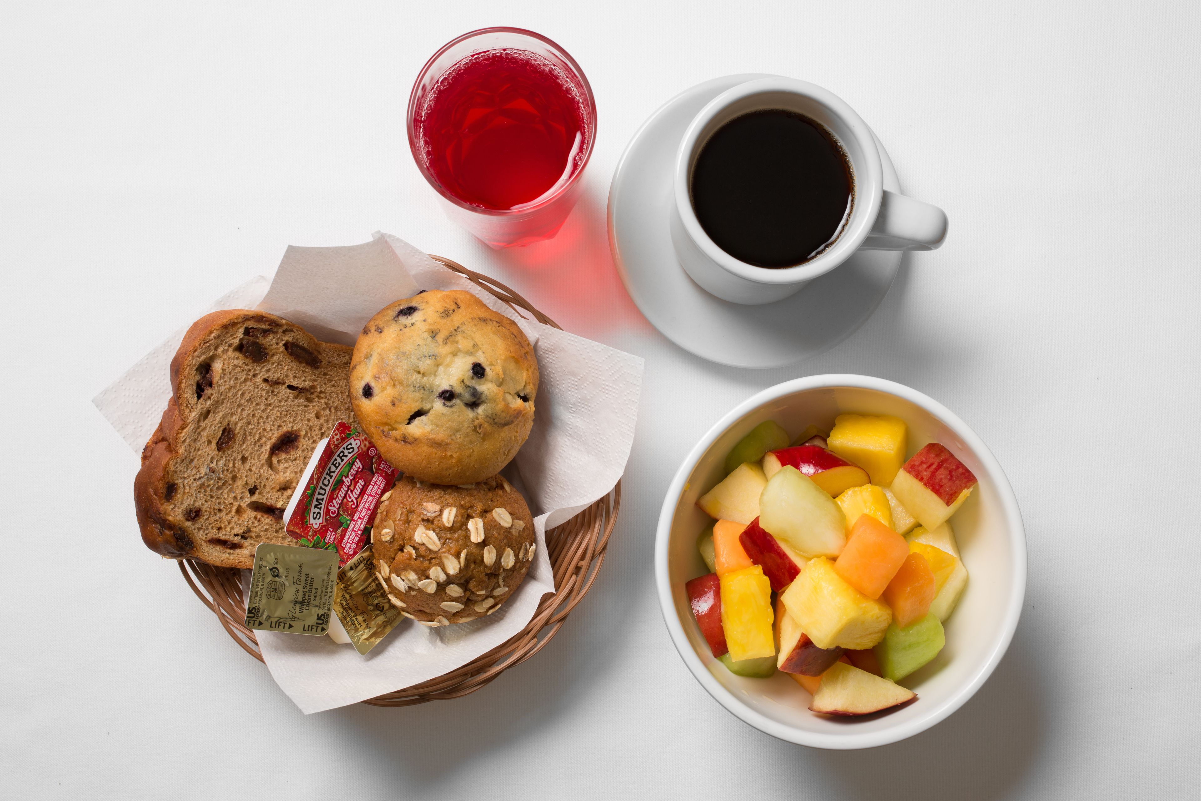 A breakfast setting with a basket of muffins, a glass of red juice, a cup of black coffee, and a bowl of fruit.