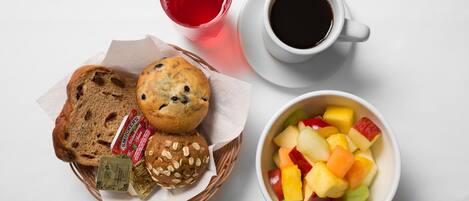 Un desayuno con una cesta de muffins, un vaso de jugo de frutas rojas, una taza de café negro y un tazón de fruta.