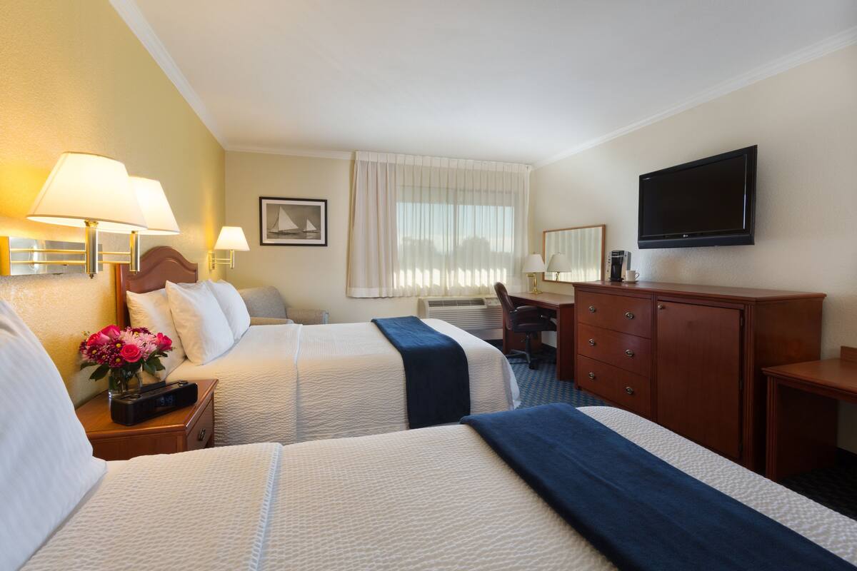 A hotel room with two beds, a desk, a chair, a TV, and a nightstand with a lamp and flowers.