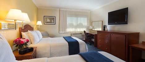 A hotel room with two beds, a desk, a chair, a TV, and a nightstand with a lamp and flowers.