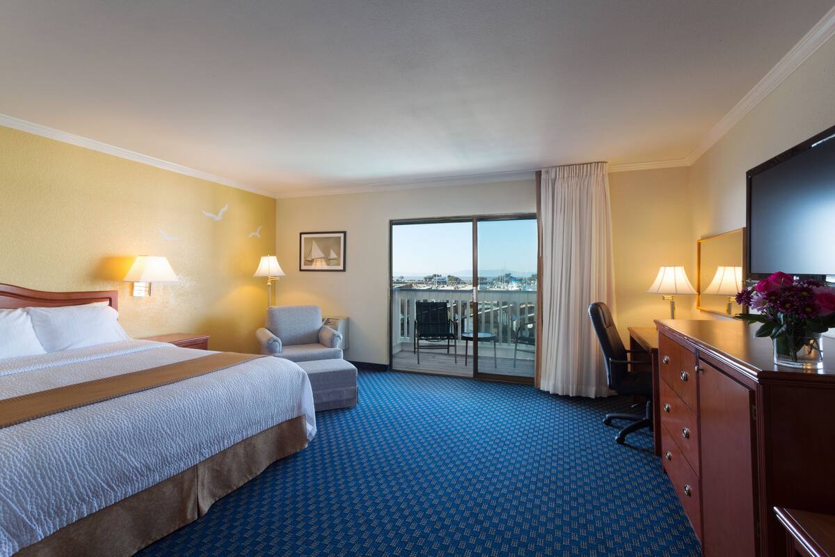 A hotel room with a large bed, a desk, a chair, a dresser, and a television. There is a balcony with a view of the city.