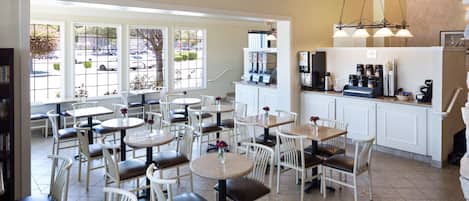 A cafe with round tables and white chairs, a coffee bar, and large windows with a view of parked cars.