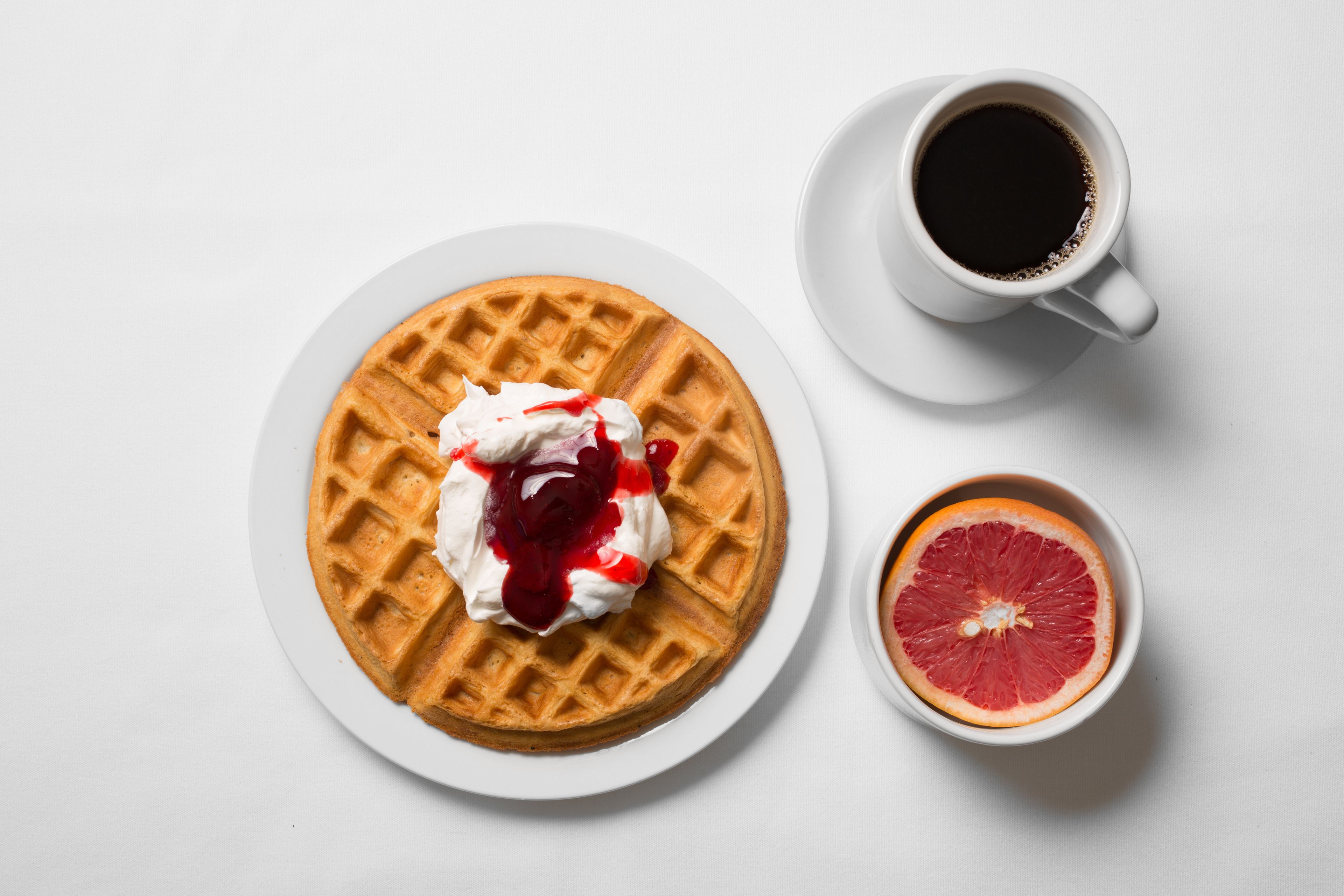 Um waffle com chantilly e molho de frutos vermelhos sobre um prato, uma chávena de café preto e uma toranja cortada ao meio sobre uma superfície branca.