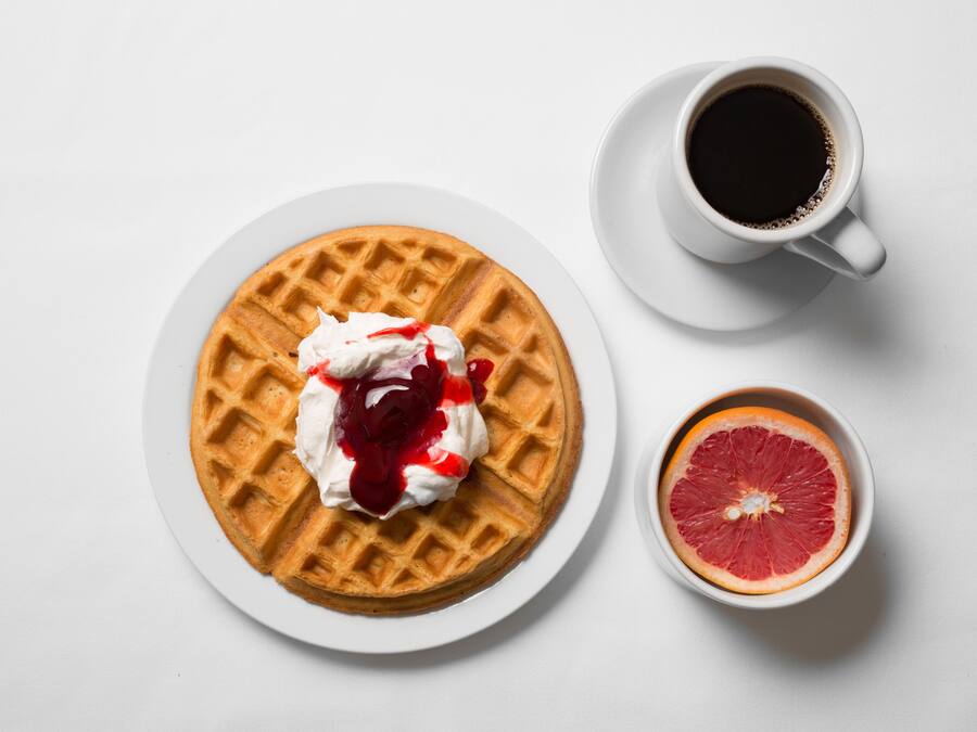 A waffle with whipped cream and berry sauce on a plate, a cup of black coffee, and a half grapefruit on a white surface.
