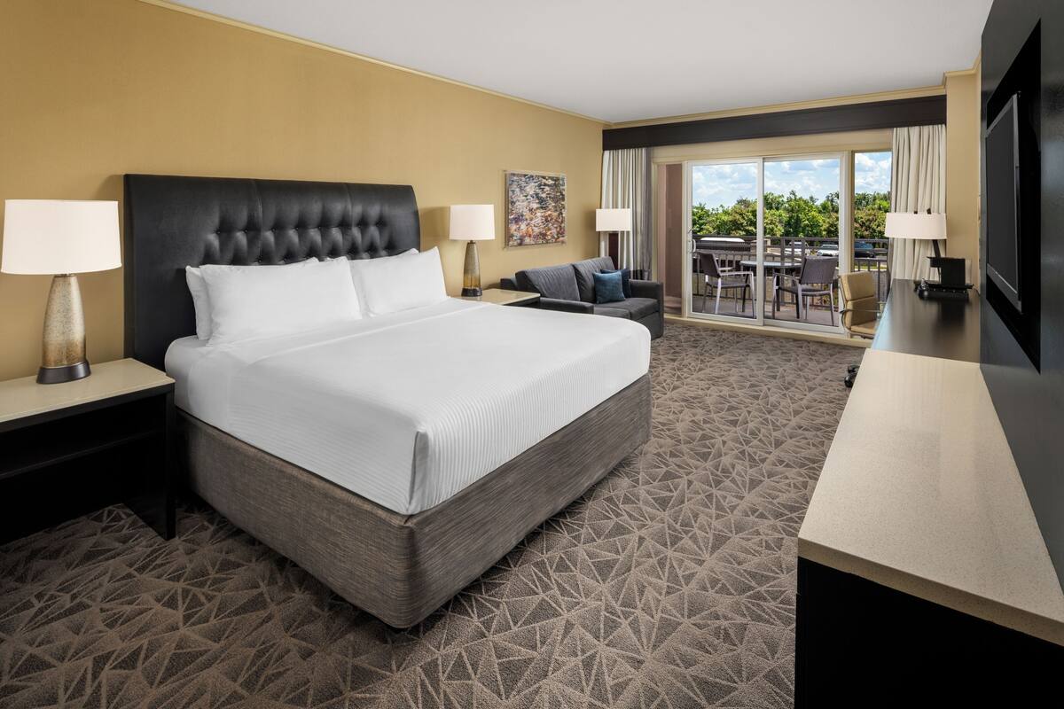 A hotel room with a large bed, two bedside tables with lamps, a sofa, a TV, and a balcony with outdoor seating.